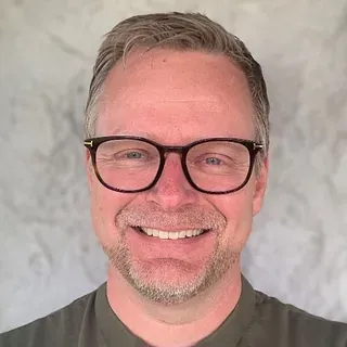 Mann med briller og kort hår smiler mot kamera foran lys bakgrunn.