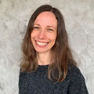 Kvinne med langt brunt hår smiler mot kamera foran lys bakgrunn.